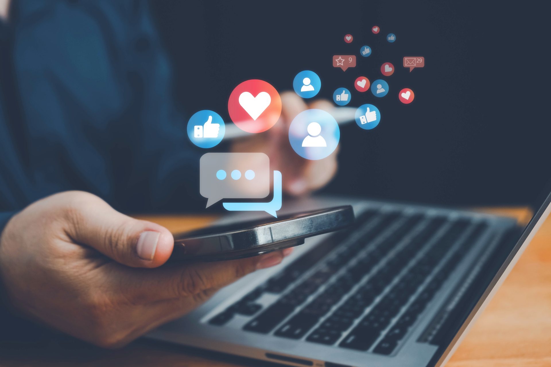 Man sitting at a laptop holding a phone, with social media icons visually emerging from the phone screen.